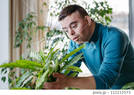 Young man with Down syndrome taking care of indoor plant, touching, snuggling plant leaf. 112215573