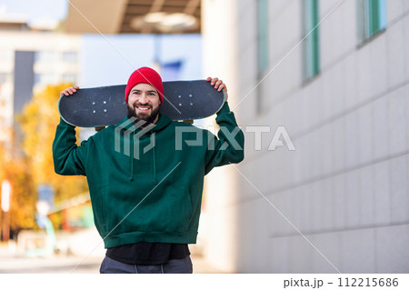 Stylish skateboarder with skateboard behind head, looking at camera. Young man skating outdoors in the city. Concept of skateboarding as sport and lifestyle. 112215686