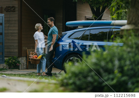 Couple standing by their electric car. Electric vehicle with charger in charging port. Couple standing by their electric car. Electric vehicle with charger in charging port. 112215698