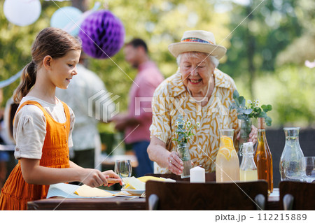 Girl helping grandmother to set outdoor table for summer garden party. Bringing plates, food, and drinks. 112215889