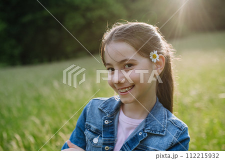 Portrait of beautiful young girl with flower behind ear, standing on meadow, enjoying warm spring day. 112215932