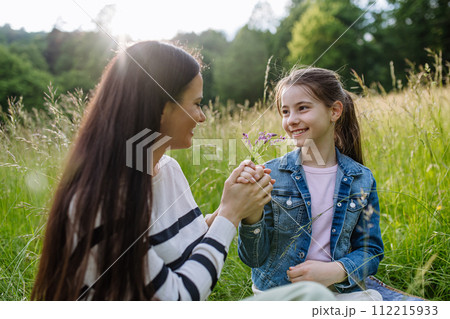 Beautiful mother with daughter, holding hands, sitting in the grass at meadow. Concept of Mother's Day and maternal love. 112215933