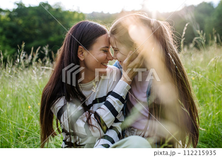 Beautiful mother with daughter, touching with foreheads, sitting in the grass at meadow. Concept of Mother's Day and maternal love. Beautiful mother with daughter, touching with foreheads, sitting in the grass at meadow. Concept of Mother's Day and maternal love. 112215935