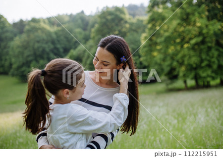 Beautiful mother with daughter, picking flowers, putting in hair, sitting in grass at meadow. Concept of Mother's Day and maternal love. 112215951