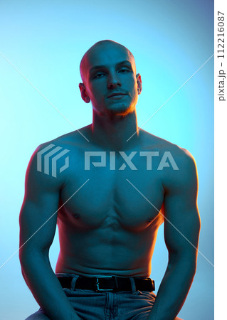 Portrait of handsome bald young man with muscular, shirtless, sportive body sitting in jeans against blue studio background in neon light 112216087