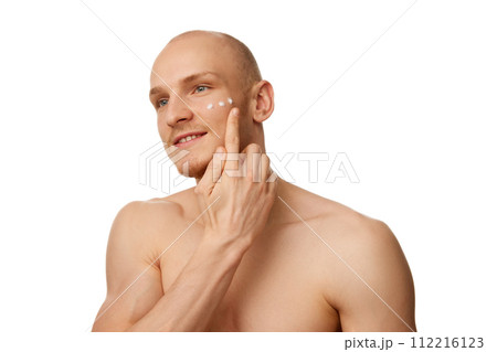 Handsome bald shirtless young man with muscular body, applying moisturizing face cream isolated against white studio background Handsome bald shirtless young man with muscular body, applying moisturizing face cream isolated against white studio background 112216123