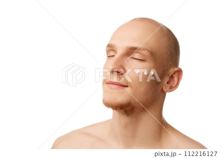 Portrait of handsome bald young man standing with eyes closed with moisturizing cream on face isolated against white studio background 112216127
