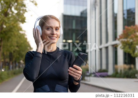 A young adult woman wearing athletic gear listens to music on her wireless headphones while holding a smartphone, evoking a sense of leisure and technology in an urban setting. 112216331