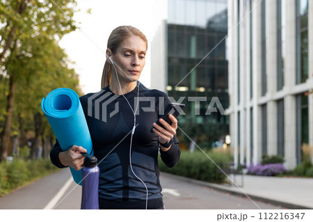 A focused female athlete carrying a yoga mat and hydration, listening to music or a podcast, and checking her smartphone before an outdoor fitness session in a city environment. 112216347