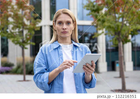 A focused professional woman stands outside on a cloudy day, using a digital tablet in front of a contemporary office building, embodying modern business and technology. A focused professional woman stands outside on a cloudy day, using a digital tablet in front of a contemporary office building, embodying modern business and technology. 112216351