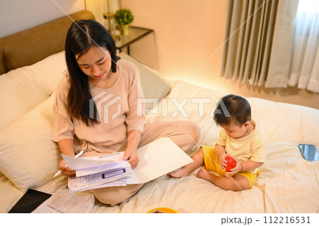 Beautiful business mom working with documents on on bed with her baby boy at home Beautiful business mom working with documents on on bed with her baby boy at home 112216531