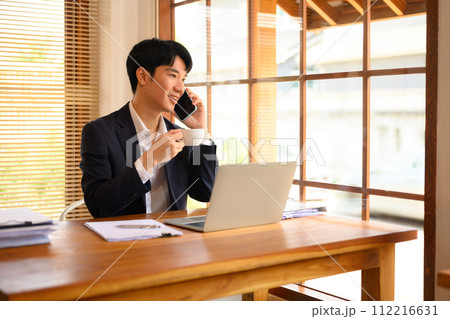 Smiling businessman drinking coffee and talking on mobile phone in office 112216631