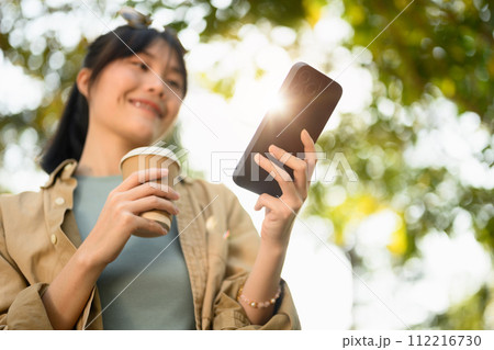 Happy young female freelancer holding paper cup and using smartphone in the summer park 112216730