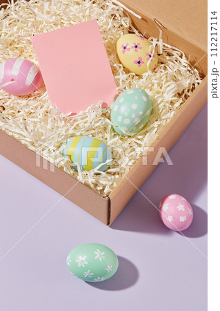 Top view of paper gift box with paper cut into shreds, colorful eggs and pink paper card decorated on purple background. Blank space for add meaningful wish. Happy Easter Top view of paper gift box with paper cut into shreds, colorful eggs and pink paper card decorated on purple background. Blank space for add meaningful wish. Happy Easter 112217114