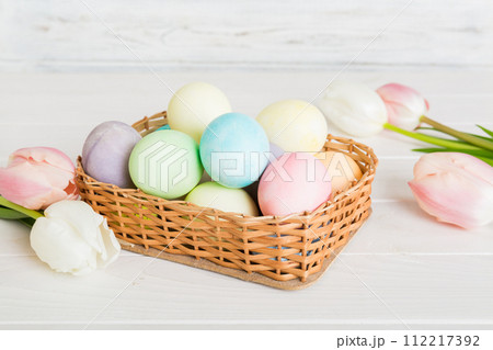 Happy Easter composition. Easter eggs in basket on colored table with yellow Tulips. Natural dyed colorful eggs background with copy space Happy Easter composition. Easter eggs in basket on colored table with yellow Tulips. Natural dyed colorful eggs background with copy space 112217392