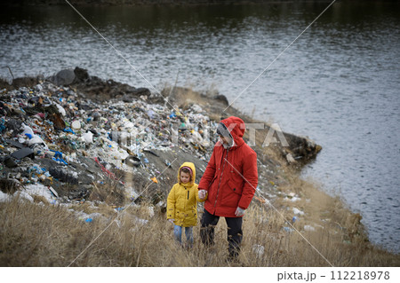 Little girl with father standing on river shore looking at piles of waste, debris on riverbank. Water and environmental pollution, eco activism concept. 112218978