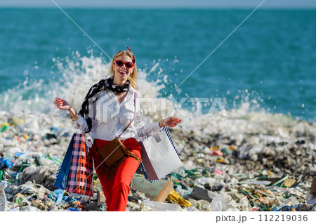 Modern woman on landfill with shopping bags. standing on pile of waste on beach. Consumerism versus pollution concept. Modern woman on landfill with shopping bags. standing on pile of waste on beach. Consumerism versus pollution concept. 112219036