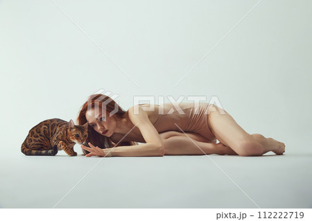 Beautiful, young, elegant redhead woman in beige bodysuit lying with purebred Bengal at against white studio background 112222719