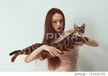 Elegant, beautiful redhead woman with bright makeup holding Bengal cat against white studio background 112222735