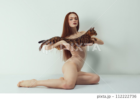 Elegant, beautiful, slim redhead woman with bright makeup holding Bengal cat against white studio background 112222736