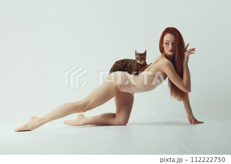 Femininity and self-identity. Passionate, beautiful young redhead woman with slim body with purebred Bengal cat lying on back against white studio background Femininity and self-identity. Passionate, beautiful young redhead woman with slim body with purebred Bengal cat lying on back against white studio background 112222750