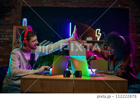 Smiling young man and African woman giving high five, sitting at table with microphones and laptop, recording online social media show 112222878