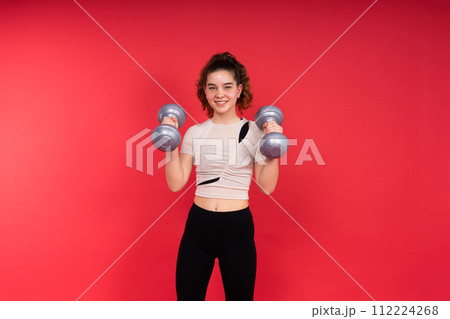 Teenage sportive girl exercises with dumbbells to develop muscles isolated on red background. 112224268