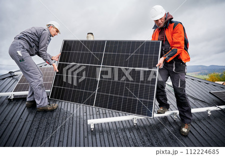 Engineers building solar panel system on roof of house. Men workers in helmets carrying photovoltaic solar module outdoors. Concept of alternative and renewable energy. 112224685