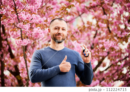 Man allergic using medical nasal drops, suffering from seasonal allergy at spring in blossoming garden. Portrait of smiling man showing thumbs up near blooming tree outdoors. Spring allergy concept. Man allergic using medical nasal drops, suffering from seasonal allergy at spring in blossoming garden. Portrait of smiling man showing thumbs up near blooming tree outdoors. Spring allergy concept. 112224715