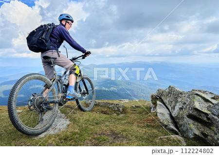 Cyclist man riding electric bike outdoors. Male tourist resting on the top of hill, enjoying beautiful mountain landscape, wearing helmet and backpack. Concept of active leisure. Cyclist man riding electric bike outdoors. Male tourist resting on the top of hill, enjoying beautiful mountain landscape, wearing helmet and backpack. Concept of active leisure. 112224802