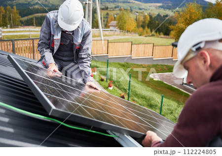 Technicians building photovoltaic solar module station on roof of house. Men engineers in helmets installing solar panel system outdoors. Concept of alternative and renewable energy. Technicians building photovoltaic solar module station on roof of house. Men engineers in helmets installing solar panel system outdoors. Concept of alternative and renewable energy. 112224805