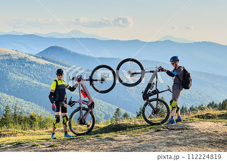 Two cyclists men riding electric bikes outdoors. Male tourists resting on the top of hill, enjoying beautiful mountain landscape, wearing helmet and backpack. Concept of active leisure. Two cyclists men riding electric bikes outdoors. Male tourists resting on the top of hill, enjoying beautiful mountain landscape, wearing helmet and backpack. Concept of active leisure. 112224819