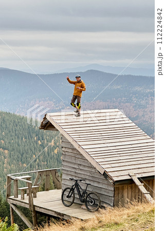 Person resting on edge of steeply sloped wooden rooftop in mountainous area after riding on electric mountain bike on cloudy day. Below the rooftop, there is wooden deck with mountain bicycle. 112224842