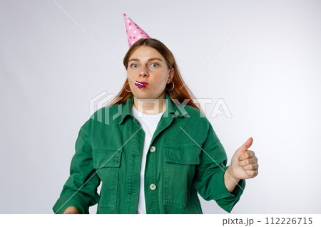 Young happy woman in birthday party hat with playing pipe showing thumbs up gesture. Emotional, young face. Human emotions, facial expression and holiday concept. Young happy woman in birthday party hat with playing pipe showing thumbs up gesture. Emotional, young face. Human emotions, facial expression and holiday concept. 112226715