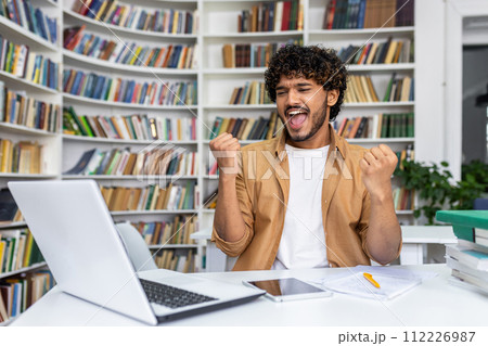 Joyful moment captured as a young professional exults in success at their desk, surrounded by a laptop, notes, and a library background, portraying a successful work accomplishment in office setting. Joyful moment captured as a young professional exults in success at their desk, surrounded by a laptop, notes, and a library background, portraying a successful work accomplishment in office setting. 112226987