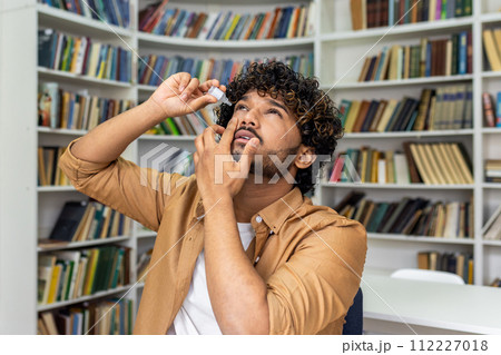 A young adult male is captured applying eye drops while standing in a cozy home library. The image reflects self-care and health management against a backdrop of bookshelves filled with books. A young adult male is captured applying eye drops while standing in a cozy home library. The image reflects self-care and health management against a backdrop of bookshelves filled with books. 112227018