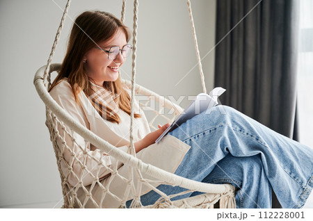 Smiling girl relaxing and reading book on indoor swing. Pretty female excited about story she reading. Cozy afternoon with book in hanging chair in home. Smiling girl relaxing and reading book on indoor swing. Pretty female excited about story she reading. Cozy afternoon with book in hanging chair in home. 112228001