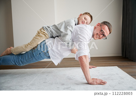 Energetic father and his little son training at home. Kid climbed dad's back while he doing plank. Young family enjoying activity games at home. 112228003