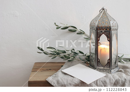 Ramadan Kareem holiday still life. Ornamental silver Moroccan lantern with olive tree branches. Blank greeting card, invitation mockup on wooden table, bench. Blurred white wall background. Ramadan Kareem holiday still life. Ornamental silver Moroccan lantern with olive tree branches. Blank greeting card, invitation mockup on wooden table, bench. Blurred white wall background. 112228843