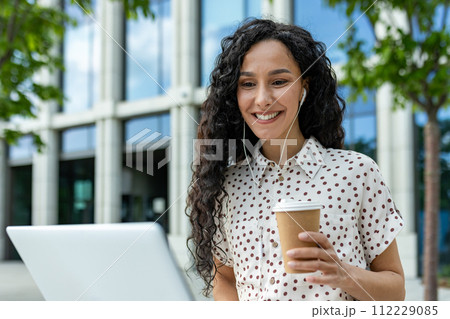 A young professional woman takes a relaxing coffee break outdoors, using her laptop against the backdrop of a modern office building. This candid moment captures the balance between work 112229085