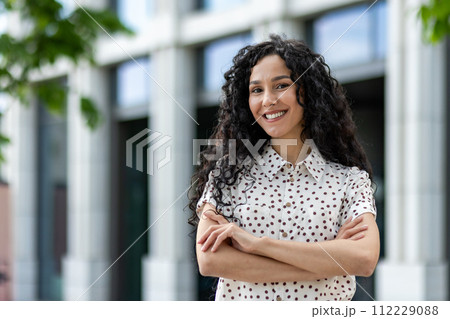 A professional businesswoman stands confidently with her arms crossed against a corporate building backdrop, exuding strength, leadership, and success. 112229088