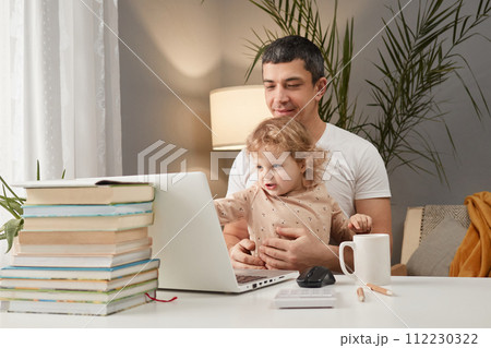 Smiling cheerful Caucasian man wearing casual clothing sitting at table in front of laptop with his infant baby daughter watching cartoons online together in home interior 112230322