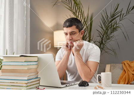 Bored sad Caucasian adult man wearing white T-shirt working at home with laptop looking at computer display with negative feeling has no mood and desire to work 112230344
