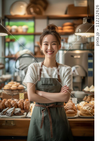 Asian woman wearing an apron with arms crossed inside a bakery shop. Asian woman wearing an apron with arms crossed inside a bakery shop. 112232626