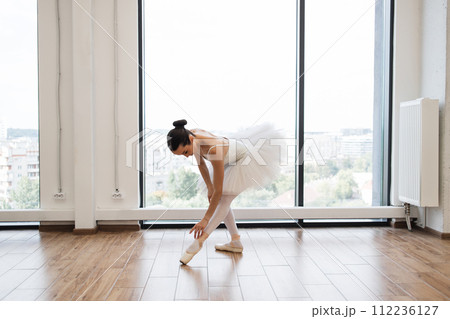 Ballerina stretching to feet while standing in dance studio with large windows. Ballerina stretching to feet while standing in dance studio with large windows. 112236127