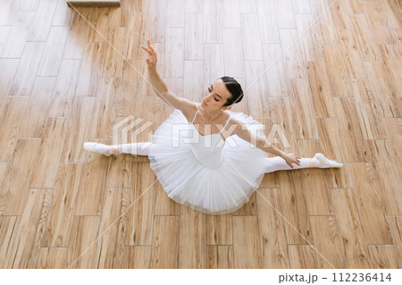 Young woman ballerina in white pack posing on background of wooden floor. Young woman ballerina in white pack posing on background of wooden floor. 112236414