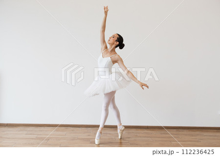 Caucasian woman ballerina in white tutu, dancing on pointe with arms overhead. 112236425
