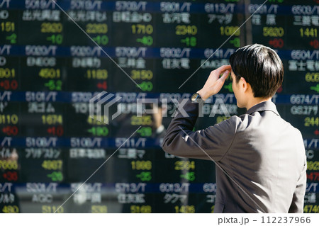 屋外の株価ボードの電光掲示板を見て不景気や株価下落に落胆し頭を抱える30代のビジネスマンの後ろ姿 112237966