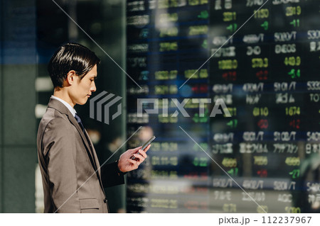 株価ボードの映る電光掲示板を前にスマートフォンで株を売買する30代のビジネスマン投資家 112237967