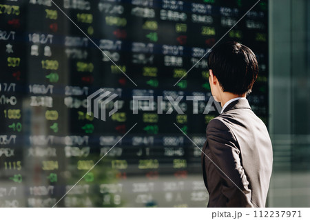 ビジネス街に設置された屋外電子掲示板の株価ボードを見る30代の日本人のビジネスマンの後姿 112237971